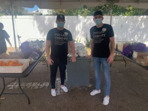 Steven and Andrew stand under a pop up tent, between two tables, in front of a cart. They both wear facemasks and their WestCMR philanthropy tee shirts.