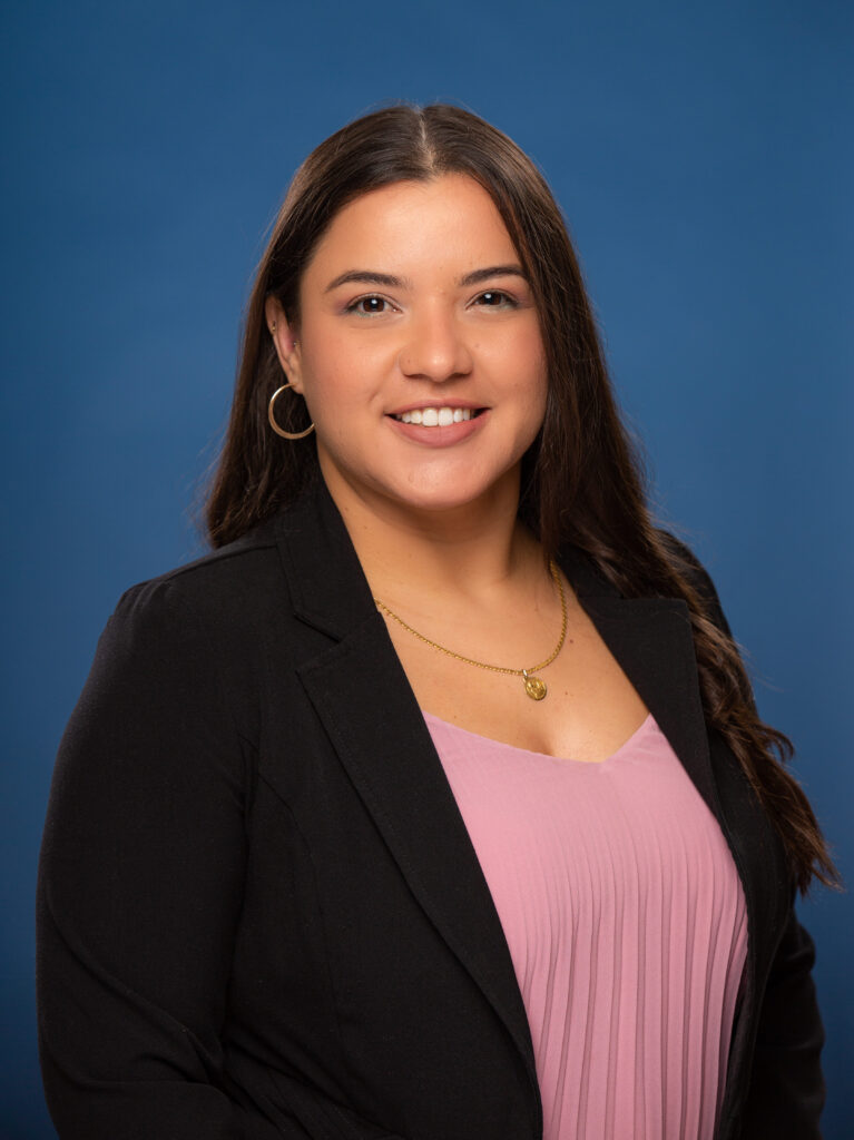 Jenny is pictured wearing a pink pleated top and black blazer. She is photographed in front of a blue background.